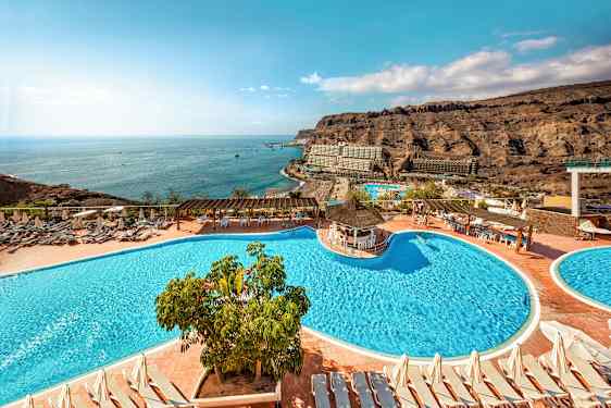 Hotelområde med swimmingpools, liggestole og udsigt over havet samt de omkringliggende klipper. (AI-genereret tekst)