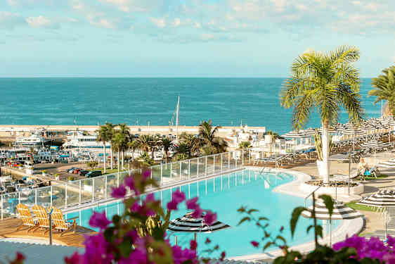 En pool med udsigt over et hav og en havn. I forgrunden er der planter og udemøbler. (AI-genereret tekst)