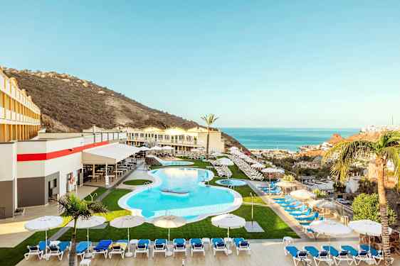 Et feriehotel med en pool, liggestole og parasoller, omgivet af et bjerglandskab og udsigt over havet. (AI-genereret tekst)