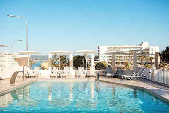 Udendørs pool med liggestole og parasoller, i baggrunden ses bygninger og havet. (AI-genereret tekst)