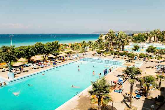 En udendørs pool omgivet af liggestole og parasoller, med palmer og havet i baggrunden. (AI-genereret tekst)