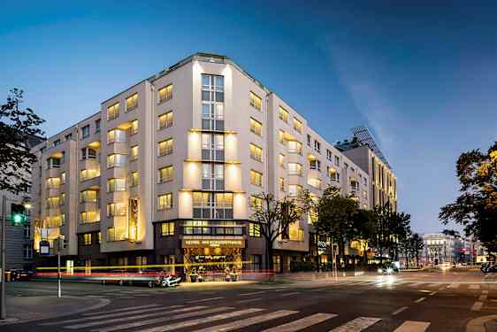 En bygning oplyst om aftenen, med flere etager og vinduer, beliggende ved et vejkryds. (AI-genereret tekst)