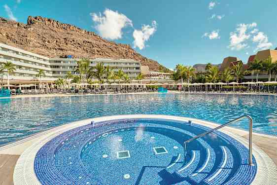 Hotellets poolområde med en mindre rund pool i forgrunden og omkringliggende bygninger samt bjerge i baggrunden. (AI-genereret tekst)