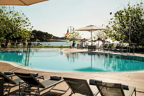 Pool med liggestole, træer og hav i baggrunden. (AI-genereret tekst)