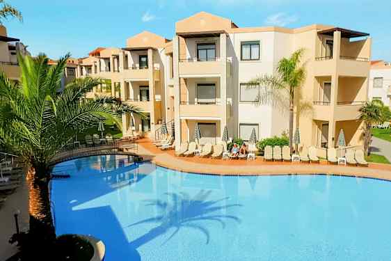 En udendørs pool omgivet af liggestole og palmer ved en beige hotelbygning. (AI-genereret tekst)