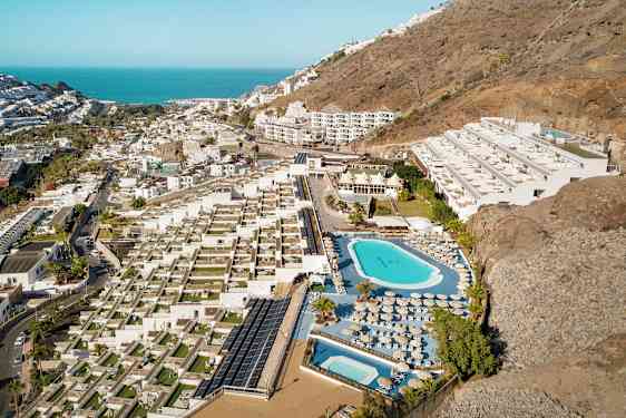 Luftfoto af et hotelområde med pools, liggestole og terrasser, beliggende ved en skråning nær kysten. (AI-genereret tekst)