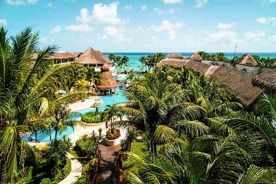 Et stort resort med pools, palmer og bygninger med stråtag, beliggende tæt på havet under en blå himmel. (AI-genereret tekst)