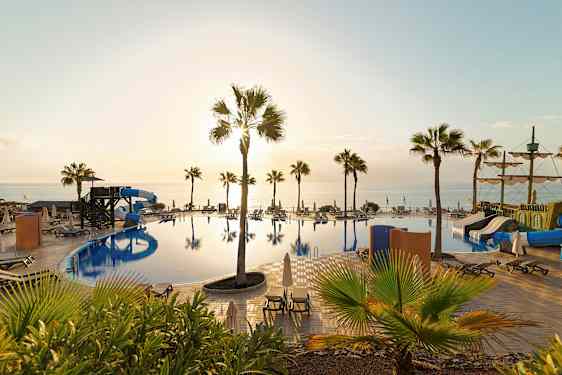 Billede af en pool med vandrutsjebaner, solsenge og palmer samt havet i baggrunden ved solnedgang. (AI-genereret tekst)