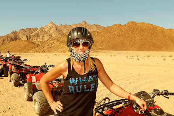 Kvinde står ved en rød quad-bike på et sandet ørkenområde med bjerge i baggrunden. (AI-genereret tekst)