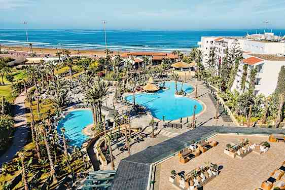 Hotelområde med pools og omkringliggende haver, beliggende tæt på en sandstrand og havet. (AI-genereret tekst)