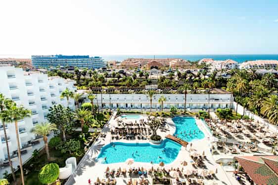 Udsigt over et hotelområde med pools, liggestole, palmer og bygninger i baggrunden. (AI-genereret tekst)