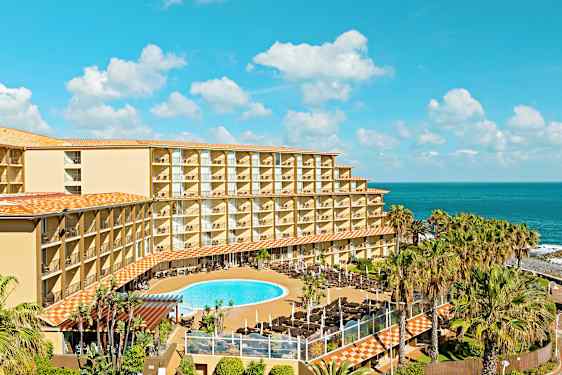 Hotelbygning med altaner, poolområde og hav i baggrunden. (AI-genereret tekst)