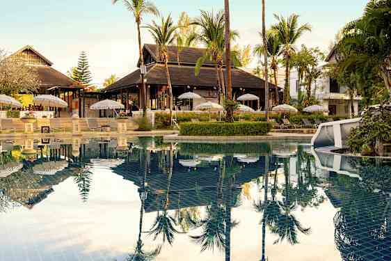 En udendørs swimmingpool med liggestole og parasoller ved siden samt omkringliggende tropisk vegetation. (AI-genereret tekst)