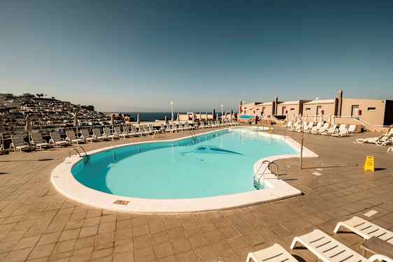 Udsigt over en udendørs swimmingpool omgivet af liggestole og parasoller. (AI-genereret tekst)