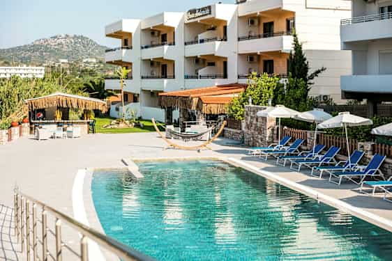 Udendørs pool med solsenge og parasoller, omkranset af bygninger på et feriested. (AI-genereret tekst)