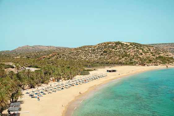 Sandstrand med turkis hav, liggestole og parasoller. Baggrund med bevoksede bjerge. (AI-genereret tekst)