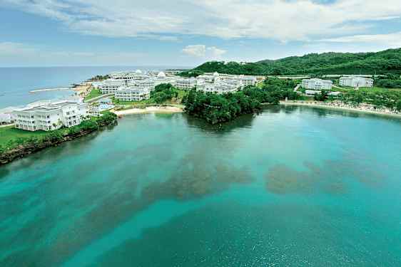 Billede af en kyst med klart vand og hvide bygninger omgivet af grønne områder. (AI-genereret tekst)