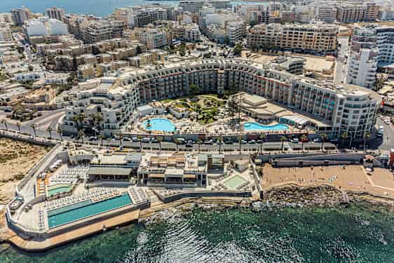 Billedet viser et omfattende feriekompleks med swimmingpools, solområder og omkringliggende bygninger tæt på kysten. (AI-genereret tekst)