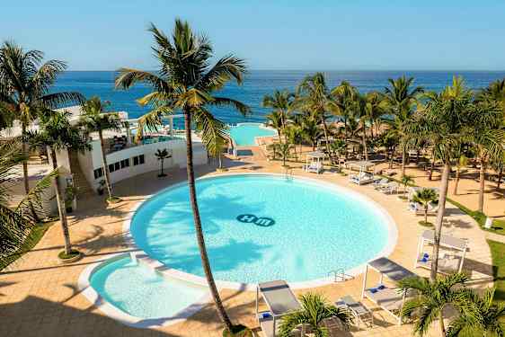 Et hotelområde med en stor rund pool omgivet af palmer, solsenge og cabanaer. Havudsigt i baggrunden. (AI-genereret tekst)