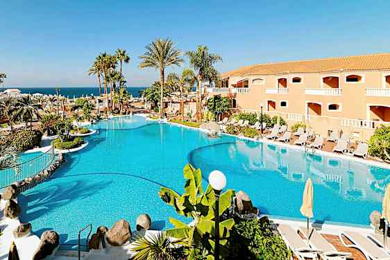 En stor pool omgivet af parasoller, liggestole, palmer og en bygning i baggrunden. (AI-genereret tekst)