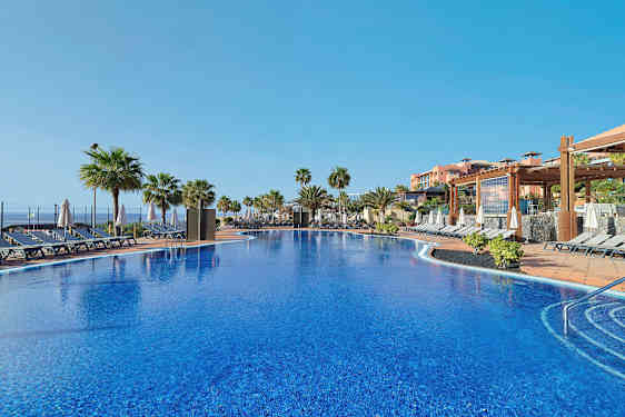 En pool med liggestole og parasoller omkring, omgivet af palmer og bygninger i baggrunden. (AI-genereret tekst)