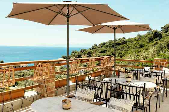 Terrasse med borde, stole og parasoller med udsigt over havet og frodige omgivelser. (AI-genereret tekst)