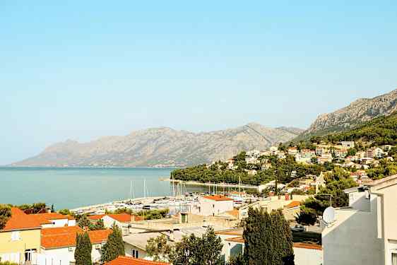 Udsigt over en kystby med røde tage, en småbådehavn og bjerge i baggrunden. (AI-genereret tekst)