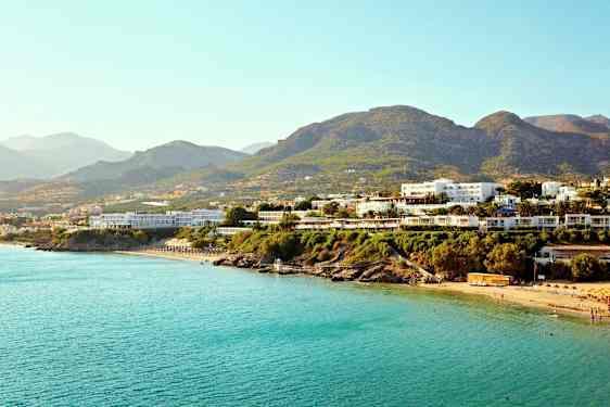 Flere bygninger nær en kyst med blåt hav og bjerge i baggrunden. (AI-genereret tekst)