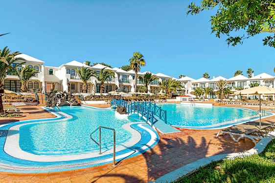 Et hotel med hvidmalede bygninger og en stor blå pool foran, omgivet af palmer og grønne områder. (AI-genereret tekst)