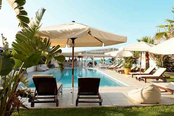 En pool omgivet af solsenge, parasoller og grønne planter. I baggrunden ses havet og en bygning. (AI-genereret tekst)