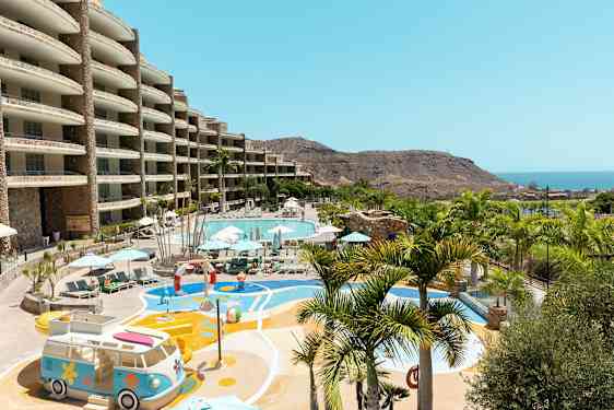 Udendørs område med en stor pool omgivet af liggestole og parasoller samt legeplads med udsigt over havet og bjerglandskab. (AI-genereret tekst)