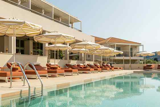 En swimmingpool omgivet af liggestole og parasoller ved en bygning. (AI-genereret tekst)