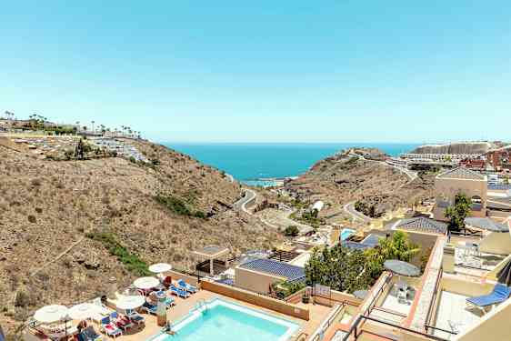 Udsigt over en pool, hotelbygninger og landskab mod havet. (AI-genereret tekst)