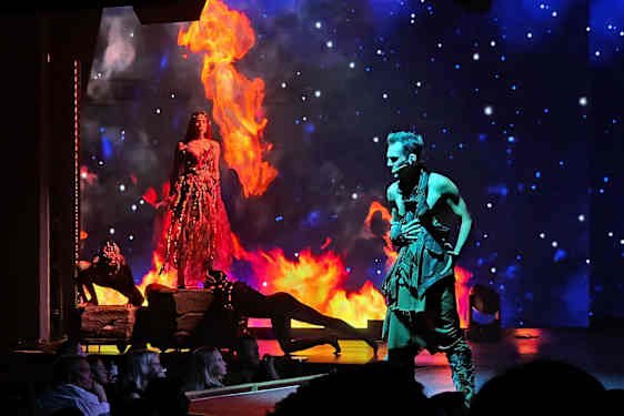 En scene med en kvinde i en ilddragt omgivet af flammer og en mand i sort dragt, med en stjernehimmel i baggrunden. (AI-genereret tekst)