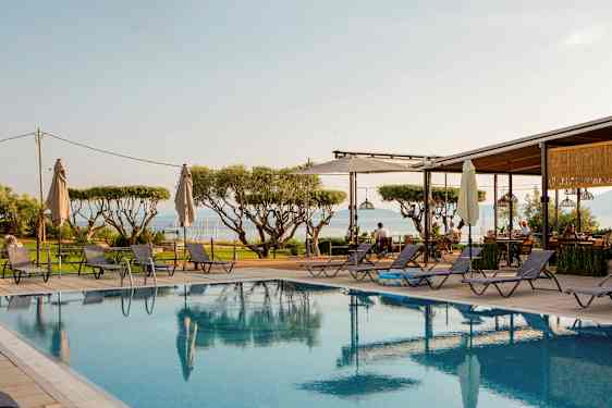 Udsigt over en pool med liggestole omkring, træer i baggrunden og havet i det fjerne. (AI-genereret tekst)