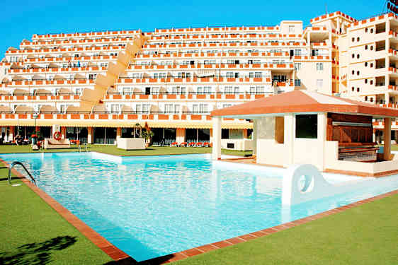 En stor pool omgivet af grønne områder og et hotel med flere etager i baggrunden. (AI-genereret tekst)