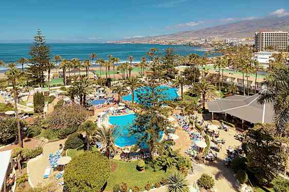 Udsigt over et ferieanlæg med pools, palmer og mennesker, der solbader og svømmer tæt på havet. (AI-genereret tekst)