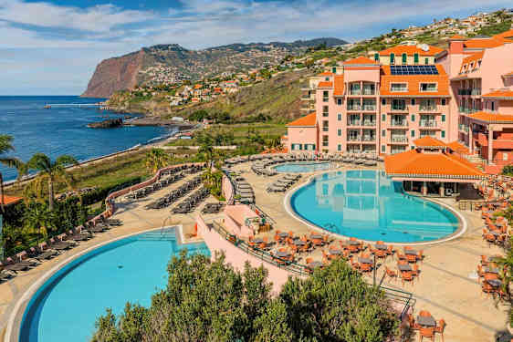 Et hotelområde ved kysten med to store pools, liggestole, borde og omkringliggende bygninger med orange tage. Baggrunden viser en klippe ved havet og boligområder langs skråningen. (AI-genereret tekst)