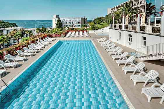 En rektangulær pool omgivet af liggestole, med havet og en bygning i baggrunden. (AI-genereret tekst)