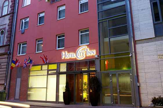 Uden for en hotelbygning med et skilt, der lyder "Hotel City Inn", og flere flag ved indgangen. (AI-genereret tekst)