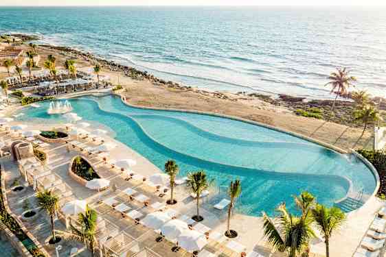 En stor pool omgivet af solsenge og parasoller ligger ved stranden med udsigt over havet. (AI-genereret tekst)