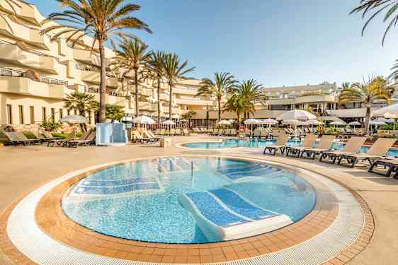 Et poolområde på et hotel med liggestole og parasoller omkring poolen, omgivet af palmer og altaner. (AI-genereret tekst)