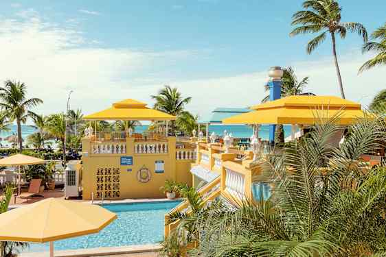 Et tropisk miljø med en swimmingpool og gule parasoller omgivet af palmer med havet i baggrunden. (AI-genereret tekst)