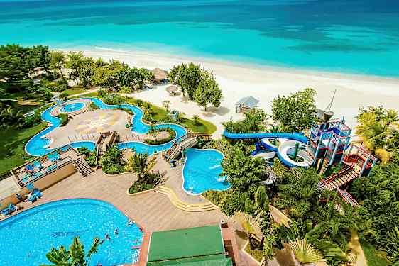 Billedet viser en strand foran et feriested med pools, vandrutsjebaner og omkringliggende vegetation. (AI-genereret tekst)