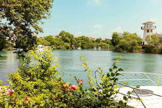 En flod med grønne buske og træer langs dens kanter. En fyrtårn-lignende bygning ses til højre. (AI-genereret tekst)