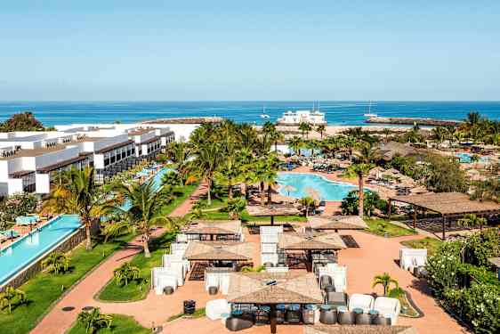 Ferieanlæg med hvide bygninger, en swimmingpool og omkringliggende grønt område, samt hav i baggrunden. (AI-genereret tekst)