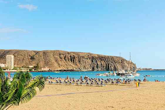 Strand med liggestole, havet og både ved en klippe. (AI-genereret tekst)