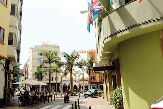 En gade med bygninger, palmer og et hotel-skilt. Folk sidder ved en café, og et motoriseret køretøj passerer. (AI-genereret tekst)