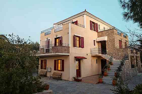 Et lyst hus med røde skodder og altaner, omgivet af grønne områder. (AI-genereret tekst)