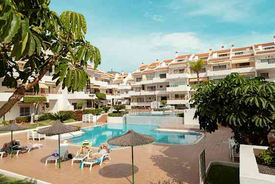 Et udendørs område med en pool, liggestole og et fleretagers hotel. Flere parasoller og grønne planter omgiver området. (AI-genereret tekst)